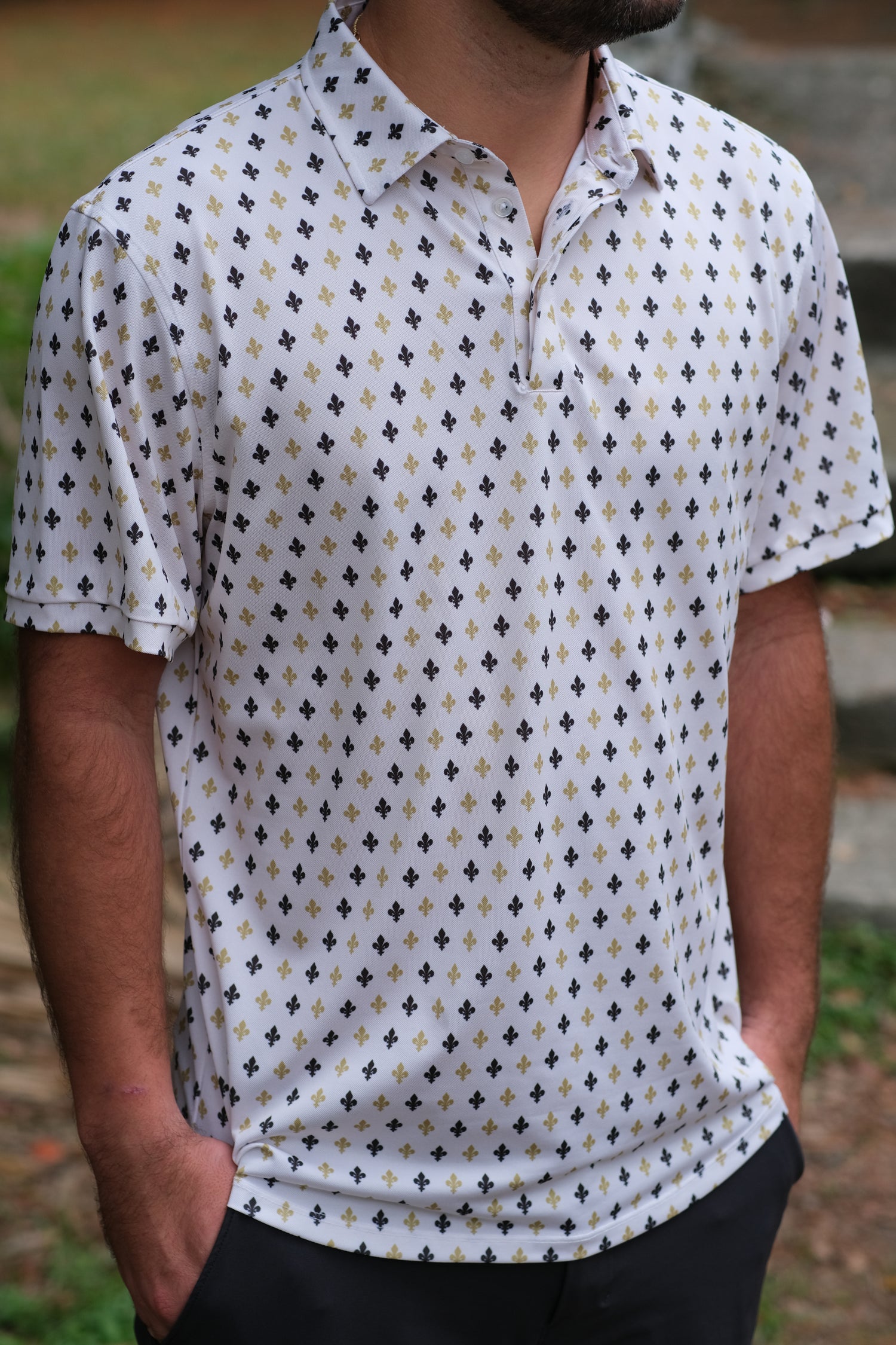 A man wearing a black and gold fleur-de-lis patterned polo shirt with a collar, standing outdoors.